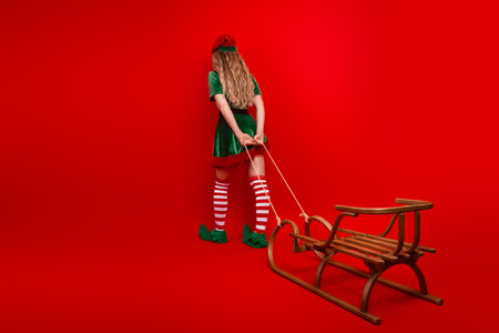 Cheerful festive elf pulling a vintage sleigh on a bright red background creating a whimsical Christmas holiday sceneの写真素材