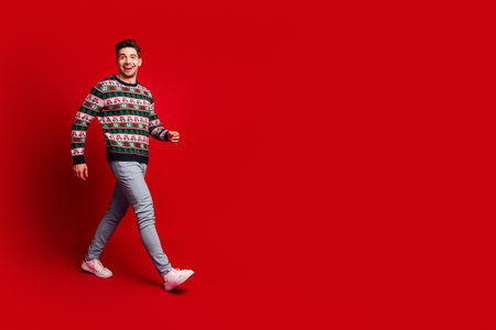 Playful man in Christmas sweater walks against bold red background sharing holiday cheer and festive styleの写真素材