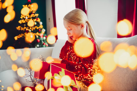 Playful Christmas moment as young girl in red sweater sits on a cozy sofa with a decorated tree and twinkling lightsの写真素材