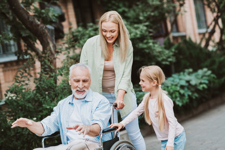 Heartwarming family care moment as grandpa in a wheelchair is guided by his daughter and granddaughter in a sunny parkの写真素材