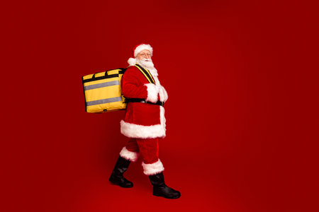Santa carries a large yellow bag and walks across a red studio background ready for Christmas celebrationの写真素材