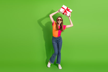 Cheerful young woman with gift box smiles and dances against bright green background in colorful...