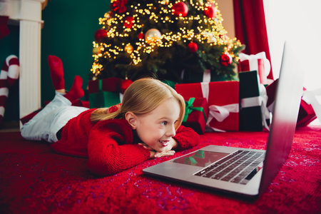 Funny cozy moment a girl lies on a red carpet at home watching a laptop beside a Christmas tree with...