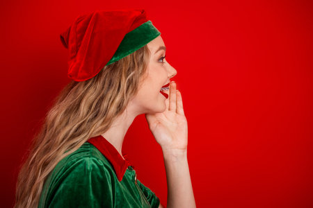 Side profile of young woman dressed as holiday elf gesturing towards red background conveying joy...