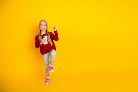 Playful young girl in red sweater holds a phone and jumps with joy against a bright yellow wall...