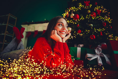 Playful girl in red sweater lies by twinkling christmas tree with gifts and warm lights creating a cozy festive scene at homeの写真素材