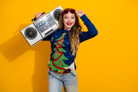 Young woman in festive Christmas sweater posing with retro boom box on vibrant yellow background, celebration moodの写真素材