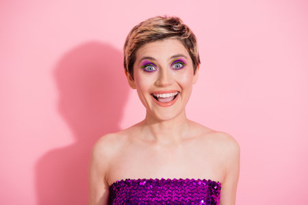 Playful young woman with short blonde hair wearing a purple sequin top stands against a pink...