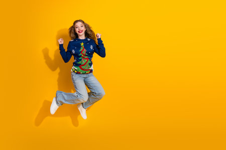 Young woman in festive Christmas sweater jumping with joy on vibrant yellow background during the...
