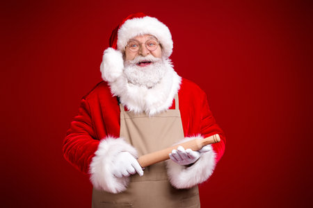 Santa in a festive red suit with a beige apron and rolling pin smiling warmly against a bright red background perfect for Christmas marketingの写真素材