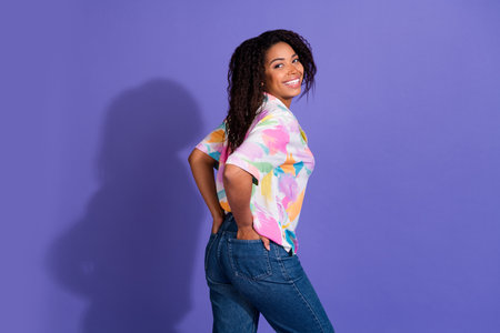 A cheerful young woman with curly black hair poses in trendy casual attire against a vibrant purple...