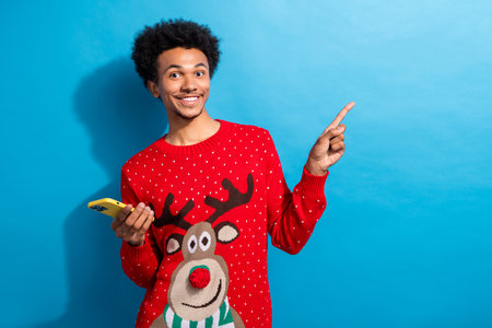Photo portrait of pretty young man hold device point empty space wear red christmas sweater masquerade theme party on blue color backgroundの写真素材