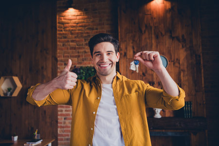 Excited young man holding keys in loft apartment, celebrating new home with thumbs upの写真素材
