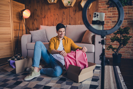 Young man enjoying weekend at home, unboxing new shirt under daylight in a cozy living room with modern interiorの写真素材