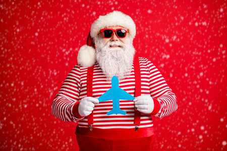 Photo of old man grey beard hold demonstrate showing blue paper plane funny wear santa claus x-mas costume suspenders sunglass striped shirt cap isolated red color backgroundの写真素材