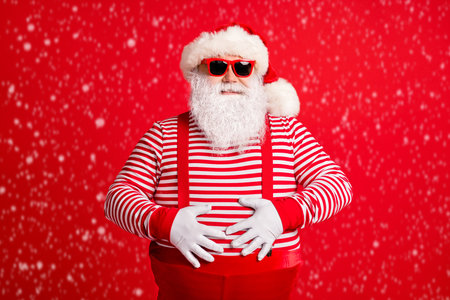 Portrait of a cheerful grey-haired Santa touching his belly, wearing gloves, isolated over a bright red background.の写真素材