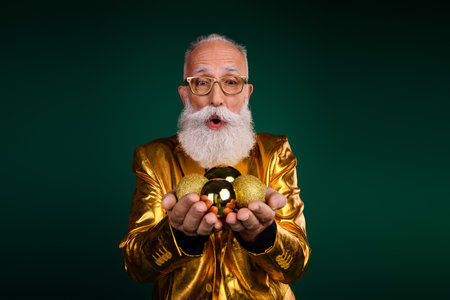 Elegant gentleman in glowing golden suit celebrates holiday with glamour, holding golden ornaments against green backgroundの写真素材