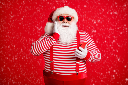 Portrait of a handsome, cheerful, amazed, bearded man dressed as Santa using a smartphone on a bright red background.の写真素材