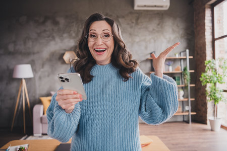 Joyful elderly woman in blue sweater using smartphone indoors at home during daylightの写真素材