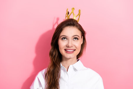 Happy young woman wearing a playful golden crown while smiling brightly against a vibrant pink backgroundの写真素材