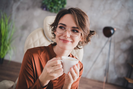 Photo of attractive young woman drink coffee espresso look empty space wear brown satin shirt modern office room interior indoors workspaceの写真素材