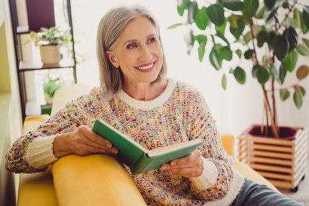 Charming elderly woman enjoying a good book in a cozy living room with daylightの写真素材