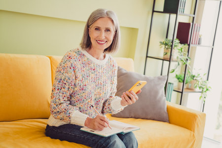 Elderly woman in a cozy autumn sweater smiling while multitasking on the couch