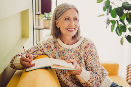 Elderly woman enjoying a peaceful afternoon writing in her journal while sitting in a comfortable living room with daylightの写真素材