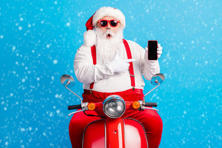 Photo of astonished grey white hair bearded santa claus x-mas christmas scooter driver rider point finger smartphone wear sunglass overalls suspenders isolated blue color backgroundの写真素材