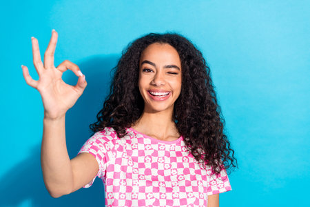 Photo of charming positive girl wear print t-shirt winking eye showing okey empty space isolated blue color backgroundの写真素材