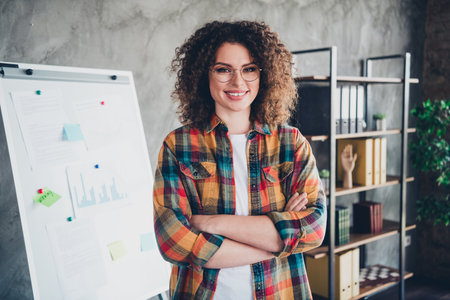 Confident businesswoman with curly hair standing in a modern office wearing a checkered shirt and glassesの写真素材