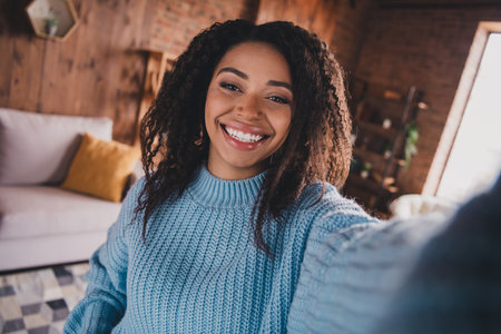 Charming young woman with afro hair smiling in a cozy living room during daylight, wearing a stylish knitted sweater.の写真素材