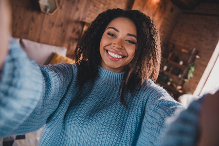 Charming young woman taking a selfie in a cozy living room with daylight, wearing a stylish knitted sweater in a casual styleの写真素材