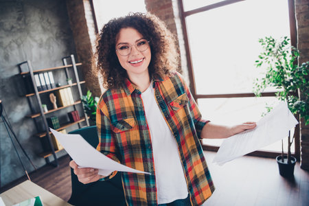 Young businesswoman with curly hair exuding confidence in a modern office while holding important paperworkの写真素材
