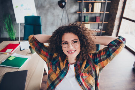 Confident young businesswoman with curly hair in casual checkered shirt relaxing in modern office interiorの写真素材
