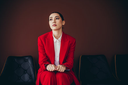 Stunning Confident Young Woman in Red Suit Sitting in Stylish Business Environmentの写真素材