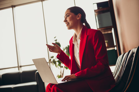 Beautiful Confident Businesswoman in Red Suit Using Laptop in Modern Office Settingの写真素材