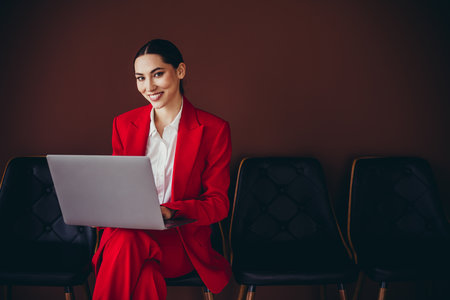 Attractive Confident Businesswoman in Red Suit Using Laptop Against Elegant Brown Backgroundの写真素材