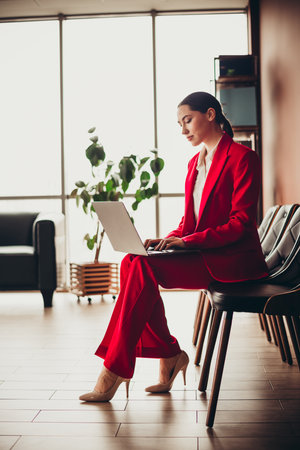 Attractive Confident Businesswoman in Red Suit Working on Laptop in Modern Office Settingの写真素材