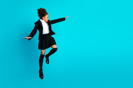 Confident young schoolgirl in stylish uniform posing joyfully against a vibrant turquoise background with an active gestureの写真素材