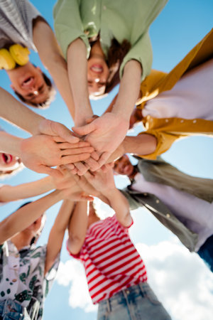 Diverse group of young best friends bonding and stacking hands outdoors on a sunny day, celebrating...