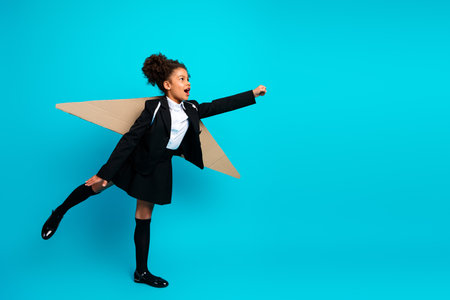 Young girl dressed in a school uniform posing playfully with a cardboard airplane on a vibrant teal backgroundの写真素材