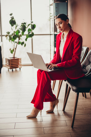 Beautiful Stunning Confident Businesswoman in a Red Suit Working on Laptop in Modern Officeの写真素材