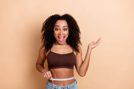 Young woman measuring her waist in a cheerful moment against a beige background wearing casual and...
