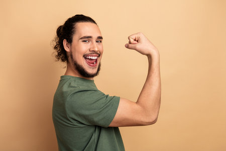 Young mixed race man flexes his bicep and smiles in a casual green t shirt against a beige...