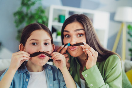 Mother and daughter having fun making funny moustaches indoors, sharing cheerful family moments at homeの写真素材