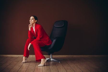 Beautiful Confident Businesswoman in Red Suit Sitting in Modern Chair Against Brown Backgroundの写真素材