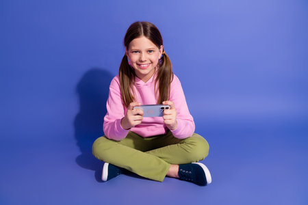 Young girl smiling and engaging with a smartphone while sitting cross-legged on a vibrant purple...