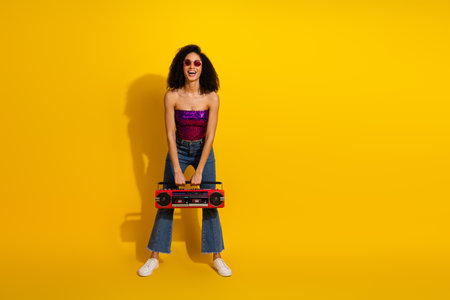 Young stylish woman wears a purple sequin top and jeans holding a red boombox smiles against a bold...