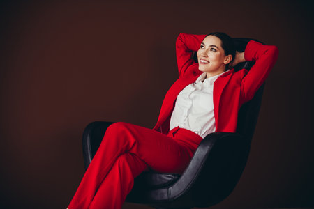 Gorgeous Confident Businesswoman in Red Suit Relaxing on Chair Against Brown Backgroundの写真素材